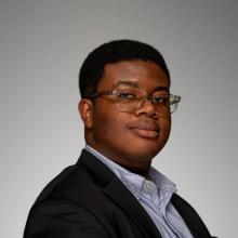 A photo of Tumi Oguntola wearing glasses and a blue and white pinstripe shirt in a suit while facing the camera at an angle.
